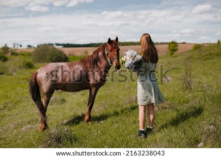 A teenage girl in a turquoise summer dress with a bouquet of lilacs looks at a brown horse in a meadow.