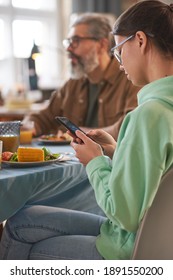 Teenage Girl Using Her Mobile Phone While Sitting At The Table With Her Family During Dinner