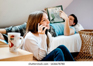 Teenage Girl using mobile phone while her friend laying on bed reading a book. Relaxing young friends at home.Brother and sister adolescence enjoy family time in room