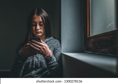 Teenage Girl Using Smart Phone In The Dark. Sad Teenager With Mobile Phone, Scared Of Threatening, Mobile Abuse. Front View Of A Sad Teen Checking Phone