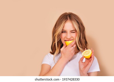 A teenage girl with wavy hair in a white t-shirt bites the lemon and grimaces in displeasure from the sour taste isolated on a beige background. Copy space. 