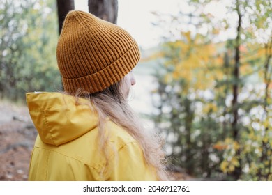 teenage girl wearing yellow raincoat and orange beanie standing in a forest and looking aside. local travel concept. fall walking alone, teen autumn leisure