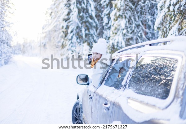 teenage girl in white sweater, vest and white
knitted hat in car window in snowy forest take selfie photo on
mobile phone, concept winter local travel during Christmas or New
Year holidays vacation