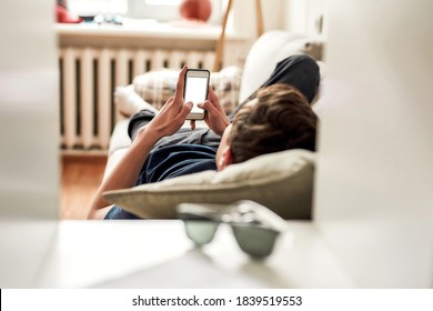 Teenage guy using his mobile phone while lying on the couch at home. The concept of smartphone addiction, apathy and frustration. Selective focus on hands and phone. Horizontal shot