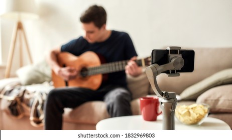 Teenage Guy Using Smartphone While Playing Guitar And Recording Video At Home. Blogging, Technology Concept. Focus On Mobile Phone On A Tripod. Web Banner