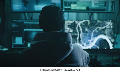 Teenage hacker with glasses watching a car on a monitor and pressing a keyboard in a dark cybercriminal hideout with lamps during a cyberattack - Powered by Shutterstock