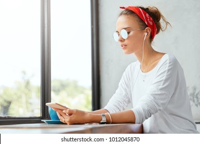 Teenage hipster girl relaxes at coffee shop with cup of espresso, listens music in playlist with earphones, sits against big window indoor, wears trendy sunglasses. Youth and technology concept - Powered by Shutterstock