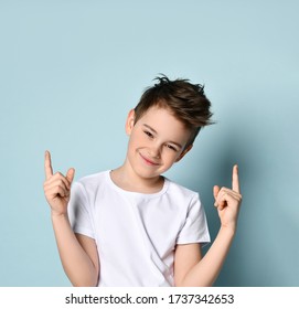 teenage kid with stylish haircut, dressed in white t-shirt. He smiling, raised his forefingers up, posing against blue studio background. Childhood, fashion, emotions. Close up, copy space
