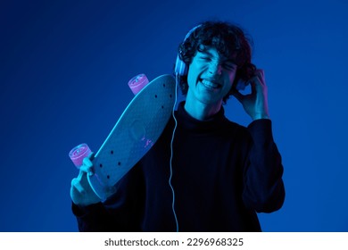 Teenage male with headphones listening to music and dancing and singing with skateboard in hand over his head, hipster lifestyle, blue background, neon light, style and trends, mixed light, copy space - Powered by Shutterstock