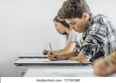 Teenage schoolboy sitting at classroom and doing exam with his classmates.