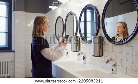 Teenage schoolgirl taking selfie in campus toilet. Side view of teen student standing in lavatory near mirror and taking selfie on smartphone