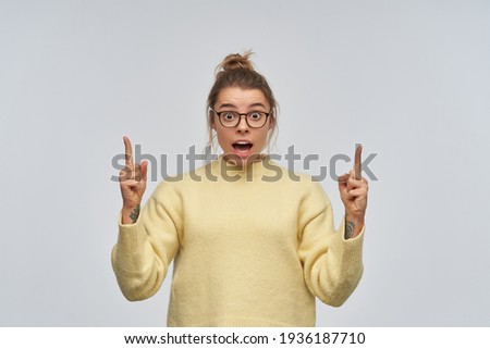 Teenage, shocked girl with blond hair gathered in bun and tattoo. Wearing yellow sweater and eyewear. Pointing index fingers up at copy space. Watching at the camera, isolated over white background