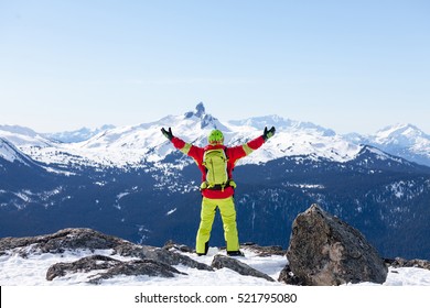 Teenage skier wearing read and green outfit with arms open wide on the top of Whistler with icon Rocky Mountains in the background.