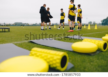 Teenage soccer players with coaches on training session. Youth team on school grass training football field. Young boys listening to coaches. Training mats, foam rollers, cones, trampoline