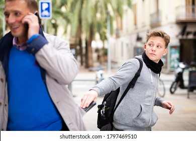 Teenage street thief stealing wallet from pocket of adult man absorbed in phone talk outdoors