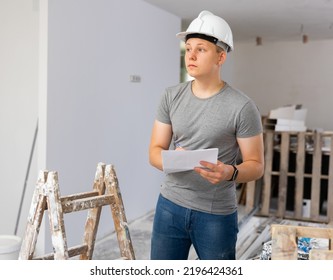 Teenage Young Man In Hardhat Doing Errand In Apartment During Repair Works, Checking Project Documentation. Teenager Learning On Part Time Work.