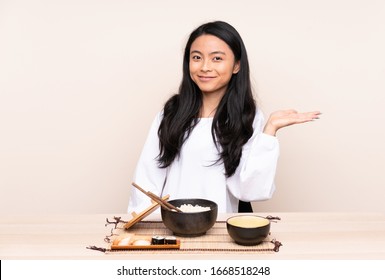 Teenager Asian Girl Eating Asian Food Isolated On Beige Background Holding Copyspace Imaginary On The Palm To Insert An Ad