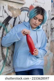 Teenager In Blue Hoodie Opening Red Water Bottle And Looking Down.Blue Haired Teen Girl Staying Outdoors Near Graffiti Wall. Clothing And Reusable Bottle Mockup.  Hipster And Adolescence Concept