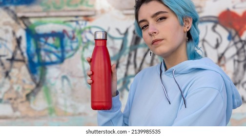 Teenager In Blue Hoodie With Red Water Bottle Looking Stright. Blue Haired Teen Girl Staying Outdoors Near Graffiti Wall. Clothing And Reusable Bottle Mockup.  Hipster And Adolescence Concept