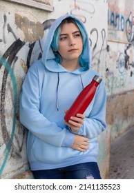 Teenager In Blue Hoodie With Red Water Bottle Looking Aside. Blue Haired Teen Girl Staying Outdoors Near Graffiti Wall. Clothing And Reusable Bottle Mockup.  Hipster And Adolescence Concept