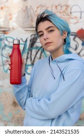 Teenager In Blue Hoodie With Red Water Bottle Looking Stright. Blue Haired Teen Girl Staying Outdoors Near Graffiti Wall. Clothing And Reusable Bottle Mockup.  Hipster And Adolescence Concept