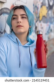 Teenager In Blue Hoodie With Red Water Bottle Looking Aside. Blue Haired Teen Girl Staying Outdoors Near Graffiti Wall. Clothing And Reusable Bottle Mockup.  Hipster And Adolescence Concept