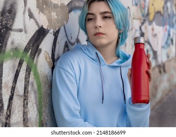 Teenager In Blue Hoodie With Red Water Bottle Looking Aside. Blue Haired Teen Girl Staying Outdoors Near Graffiti Wall. Clothing And Reusable Bottle Mockup.  Hipster And Adolescence Concept