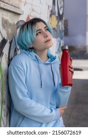 Teenager In Blue Hoodie With Red Water Bottle Looking Aside. Blue Haired Teen Girl Staying Outdoors Near Graffiti Wall. Clothing And Reusable Bottle Mockup.  Hipster And Adolescence Concept