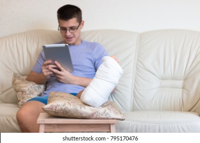 A Teenager Boy With A Broken Foot Alone At Home Doing Homework, Reading On The Tablet. Leg In A Cast. Disease. 