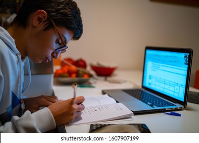 Teenager boy doing homework at home with laptop and writing notes.