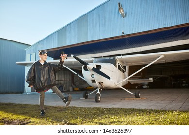 Teenager Boy In Large Pilot Jacket, Full Of Fun, Playing With Model Airplane Near Aircraft Hangar Building, Light Propeller Airplane On Background. Outdoor Shot, Full Lenght View.