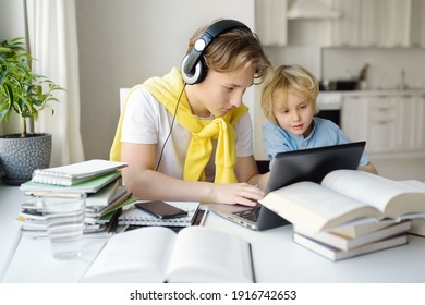 Teenager Boy Study At Home. His Younger Brother Is Playing Near. Homeschooling. Online Education And Distance Learning For Children During Lockdown. Friendship Of Siblings With A Big Age Difference.