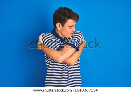 Teenager boy wearing casual t-shirt standing over blue isolated background Hugging oneself happy and positive, smiling confident. Self love and self care