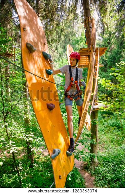 Teenager boy\
wearing safety harness passing rope bridge obstacle at a ropes\
course in outdoor treetop adventure\
park