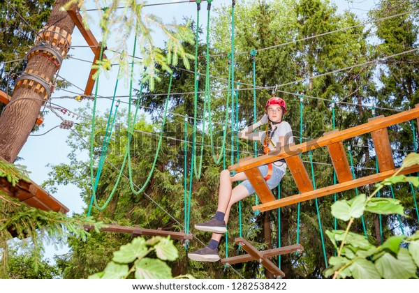 Teenager boy wearing safety harness sitting on a\
hanging rope bridge at a ropes course in outdoor treetop adventure\
park taking a break