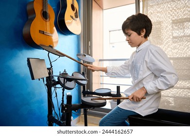 Teenager boy in white shirt and blue jeans, playing drum kit in his modern music studio room. Two acoustic electrical guitars hanging on a blue wall. People. Music. Kids hobbies and leisure activity - Powered by Shutterstock