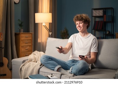 A teenager, a brunette with curly hair relaxes on the living room couch in the evening. The young boy holds the remote control in his hand and switches the channel to a funny program