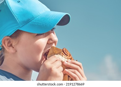 A Teenager In A Cap Eats Pastries Against A Blue Sky On A Summer Day Outside. High School Girl Eating Cinnamon Bun, Face Close Up.