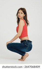 A teenager is crouching elegantly on her toes, showcasing a fashionable red crop top and fitted blue jeans. The simple white backdrop enhances her youthful spirit and relaxed pose.