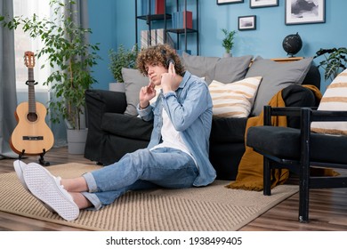 A Teenager Dressed In Jeans Pants And Shirt Is Hanging Out In The Living Room At Home. The Boy Is Watching A Concert Of His Favorite Band On TV And Listening To Their Music On Headphones.