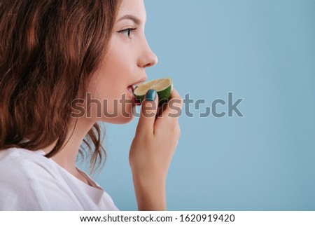 teenager eat slice of lime, make grimaces, in studio, isolated on blue background, copy space, profile view