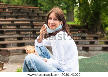 A teenager eats a sandwich outdoors between lessons. A girl wearing a medical mask from COVID-19 during a pandemic. Healthy nutrition of a schoolchild in the educational process.