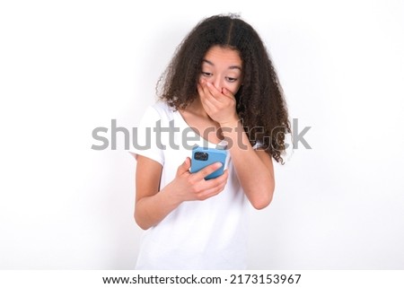 Teenager girl with afro hair style wearing white t-shirt over white background being deeply surprised, stares at smartphone display, reads shocking news on website, Omg, its horrible!