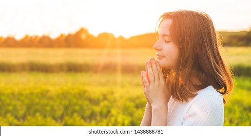 Teenager Girl closed her eyes, praying in a field during beautiful sunset. Hands folded in prayer concept for faith, spirituality and religion. Peace, hope, dreams concept