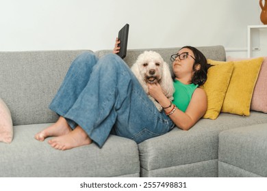 Teenager girl enjoying her free time at home, reading an e-book on a digital tablet while cuddling her pet dog on a comfortable sofa - Powered by Shutterstock