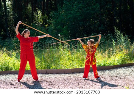 teenager girl with little boy in red sports wushu uniform are training in the park,  kung fu fighter children athletes, practicing wushu with a stick