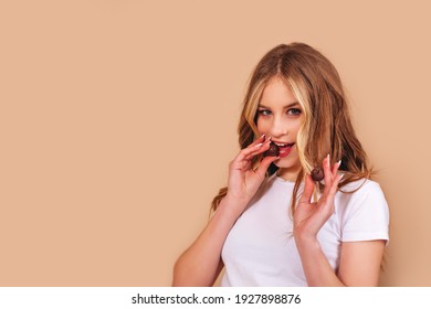 Teenager girl with long blond hair in a white t-shirt eats chocolate candies isolated on a beige background. Copy space. 