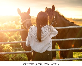 Teenager girl looking at dark horses by a metal gate to a field at stunning sunset. Warm sunshine glow. Selective focus. Light and airy soft look. Connection to nature concept. Rural area. - Powered by Shutterstock