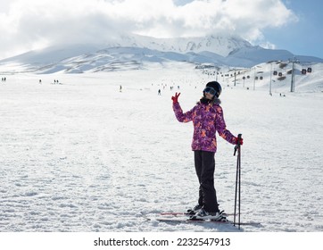 A teenager girl on skies dressed in a ski suit and helmet stands against the backdrop of snow-covered mountain ski slope and a bright blue sky. Winter. Sport and travel content   - Shutterstock ID 2232547913