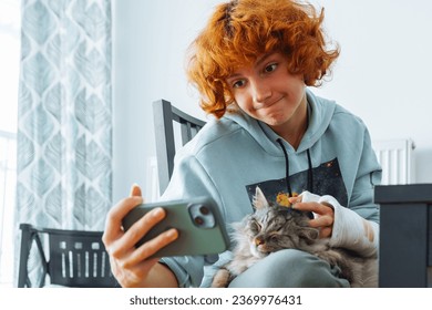 Teenager girl, red-haired, attractive, with broken arm, arm in cast, communicates using smartphone, communicates via video conference, sitting at home in living room on chair - Shutterstock ID 2369976431